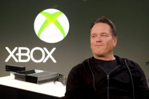 Un homme souriant devant un logo Xbox et une console, symbolisant les dix années de direction de Phil Spencer chez Xbox.