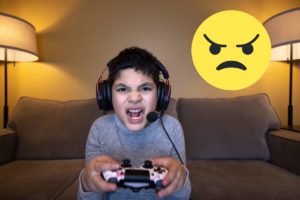Un garçon fâché jouant à un jeu vidéo avec un emoji en colère à côté.